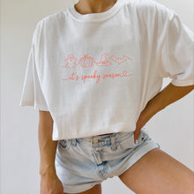 Load image into Gallery viewer, White fall graphic T-shirt with a minimalist ghost, pumpkin, witch hat, and bat design and the phrase "It's Spooky Season," paired with denim shorts for a casual autumn outfit.
