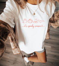 Load image into Gallery viewer, Woman wearing a white graphic T-shirt featuring a minimalist ghost, pumpkin, witch hat, and bat design with the phrase "It's Spooky Season," styled with light wash denim shorts for a casual fall outfit.

