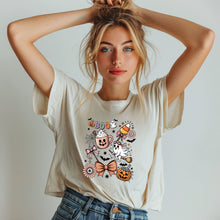 Load image into Gallery viewer, Cute Halloween graphic tee featuring a pastel ghost, pumpkin, candy corn, bow, and coffee mug design on a cream unisex T-shirt for spooky season.

